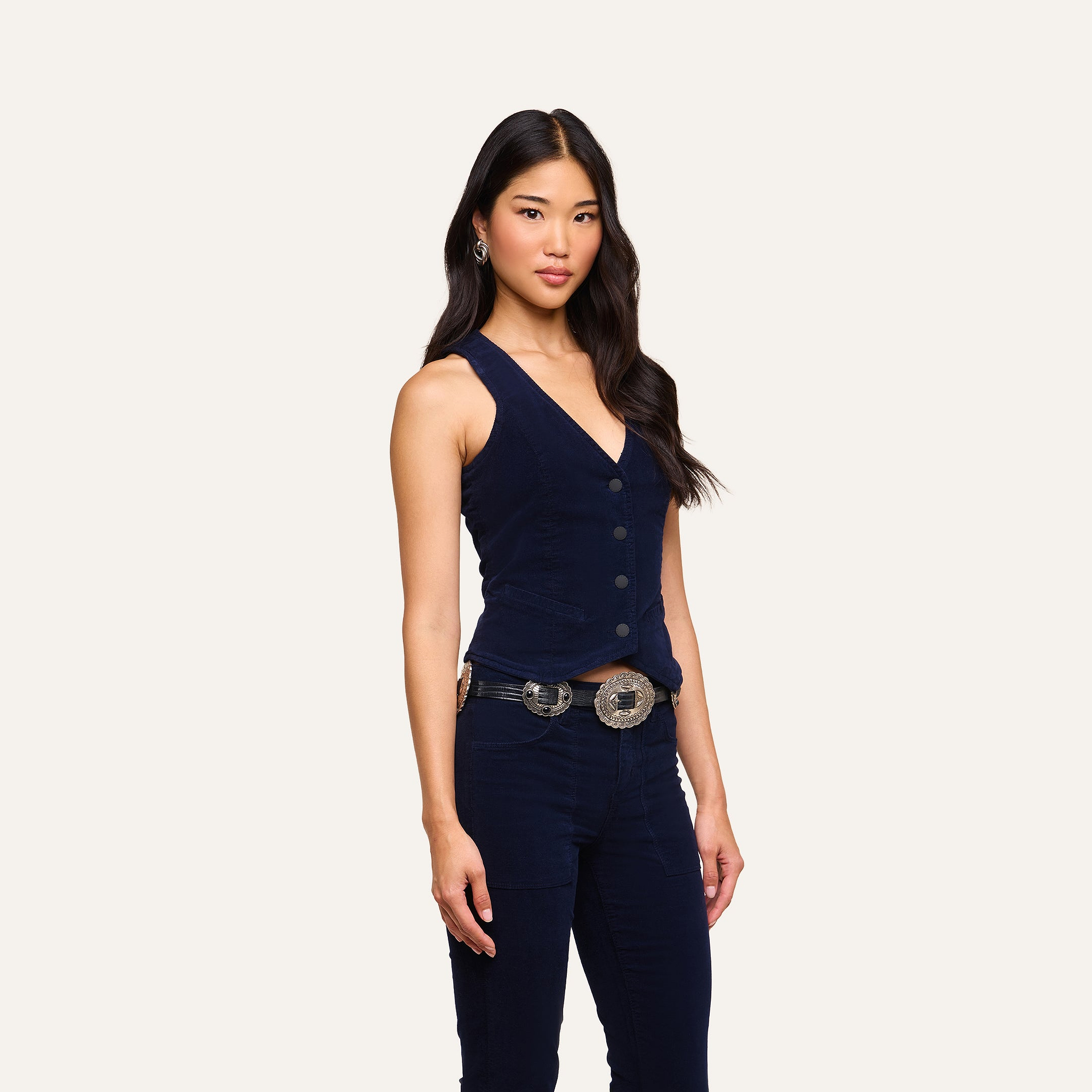 Wearing the Yelena Vest styled as a button-up top, a woman with long dark hair pairs it with matching pants and a statement belt featuring large metal buckles, all set against a simple light background.