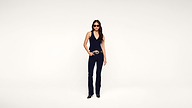 A woman in sunglasses wears a navy halter jumpsuit with a wide belt and black shoes, standing against a plain white background.