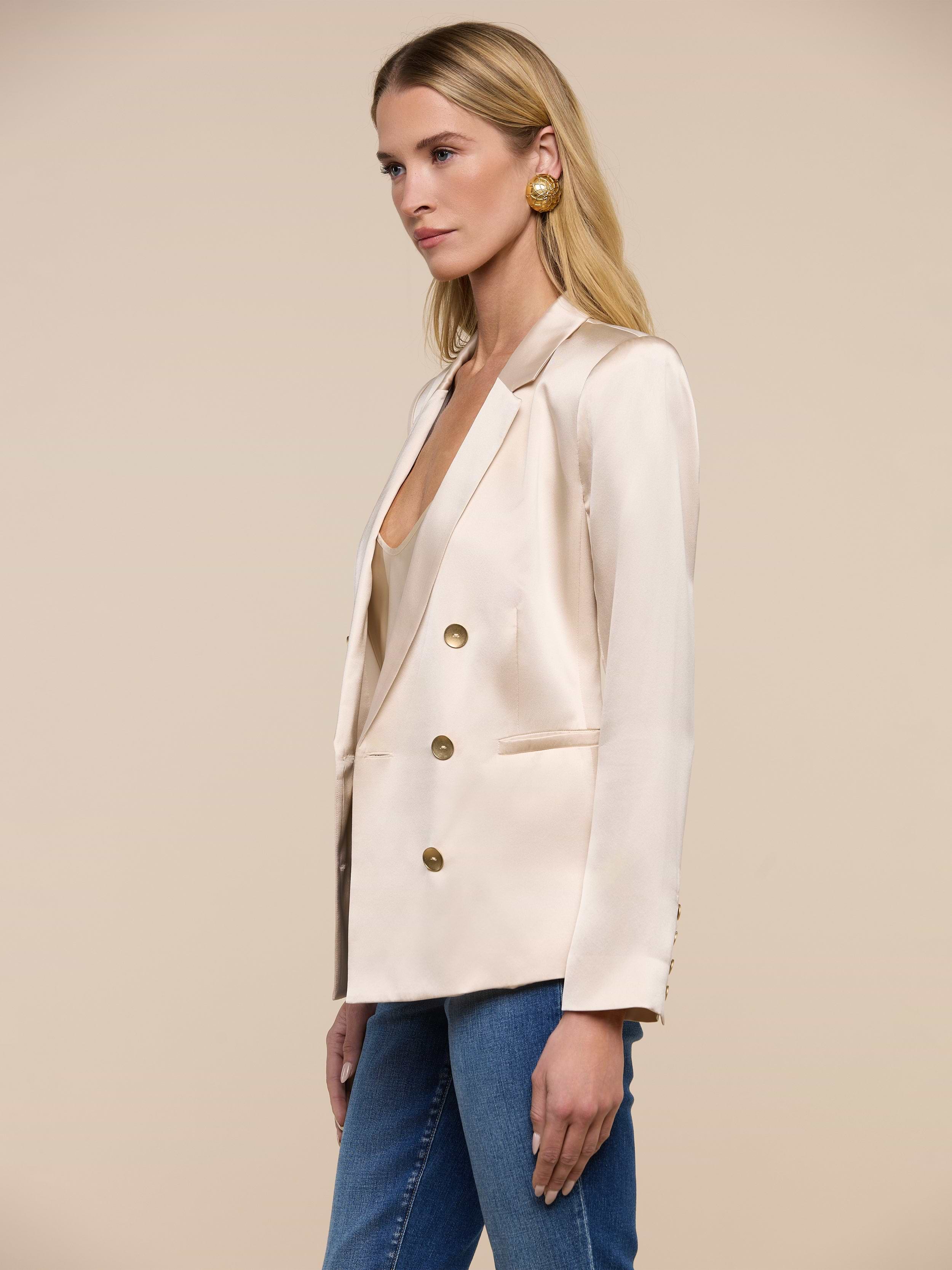 Wearing the Colin Silk Blazer—a double-breasted, cream-colored piece with gold buttons—a woman stands against a plain white background. She pairs the blazer with blue jeans and large gold earrings.