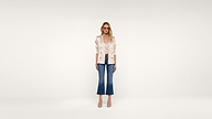 A woman stands against a plain white background, wearing a light-colored blazer, blue cropped jeans, and beige heels. She is looking forward with a neutral expression, arms relaxed at her sides.