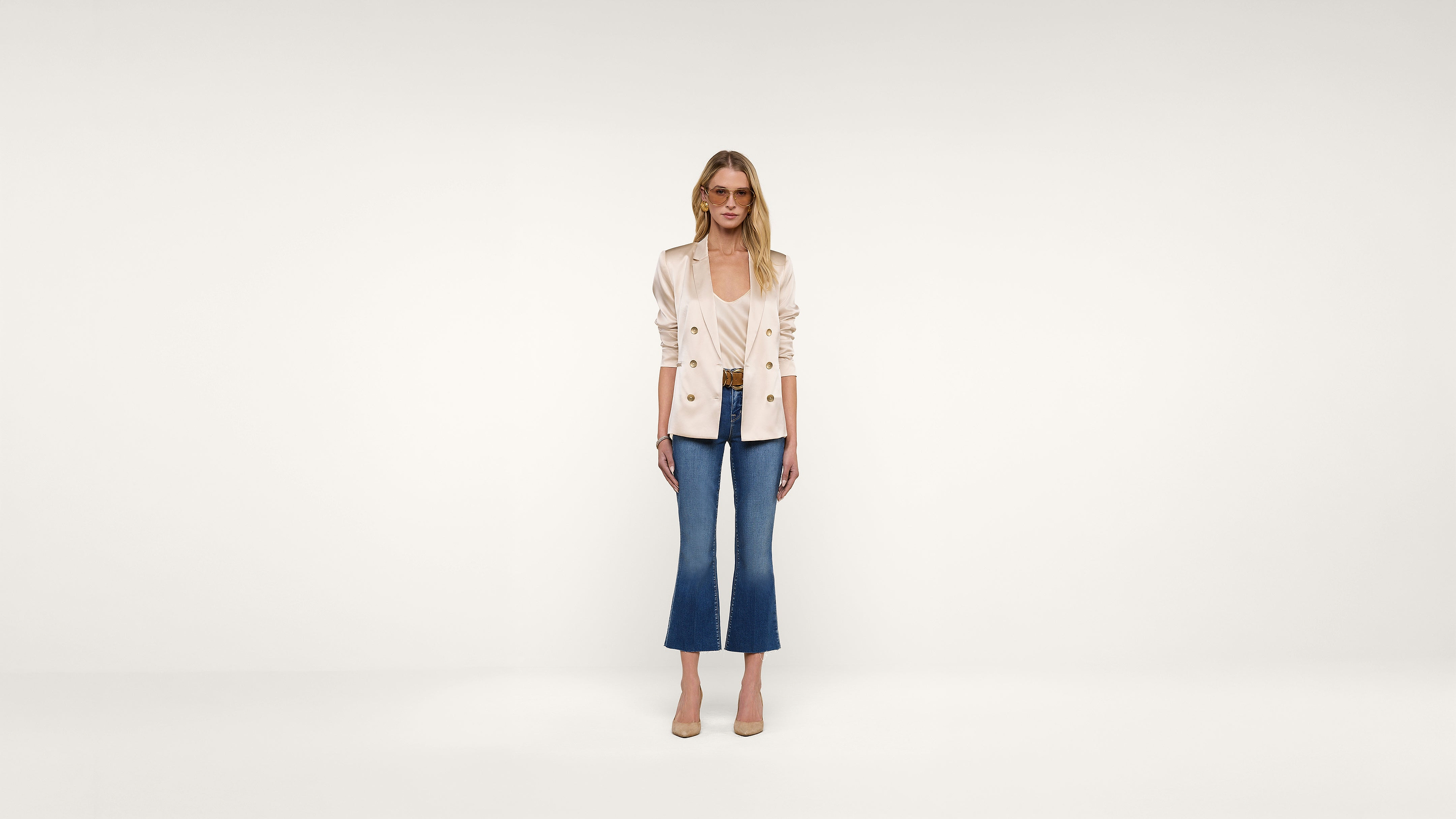 A woman stands against a plain white background, wearing a light-colored blazer, blue cropped jeans, and beige heels. She is looking forward with a neutral expression, arms relaxed at her sides.