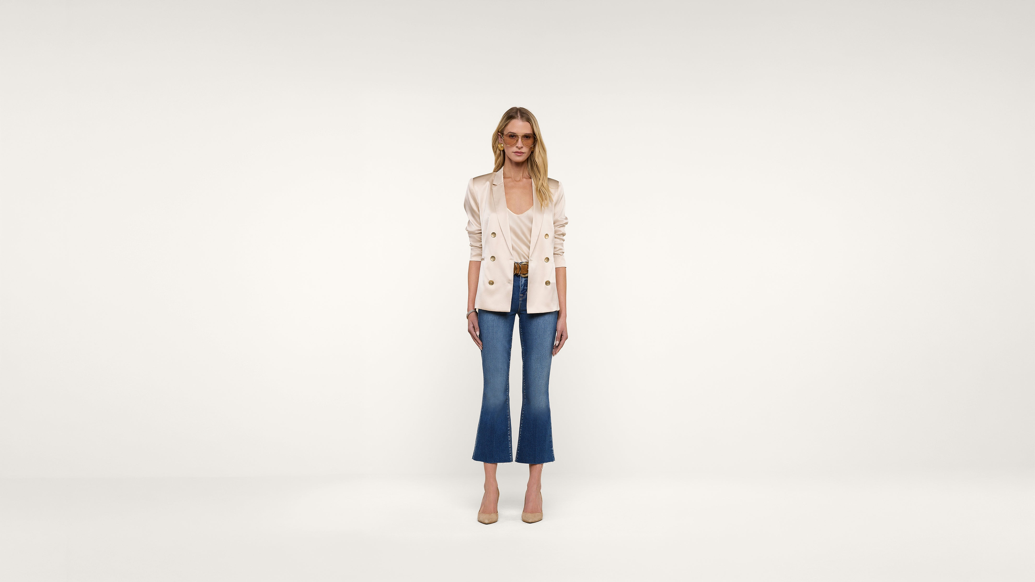 A woman stands against a plain white background, wearing a light-colored blazer, blue cropped jeans, and beige heels. She is looking forward with a neutral expression, arms relaxed at her sides.