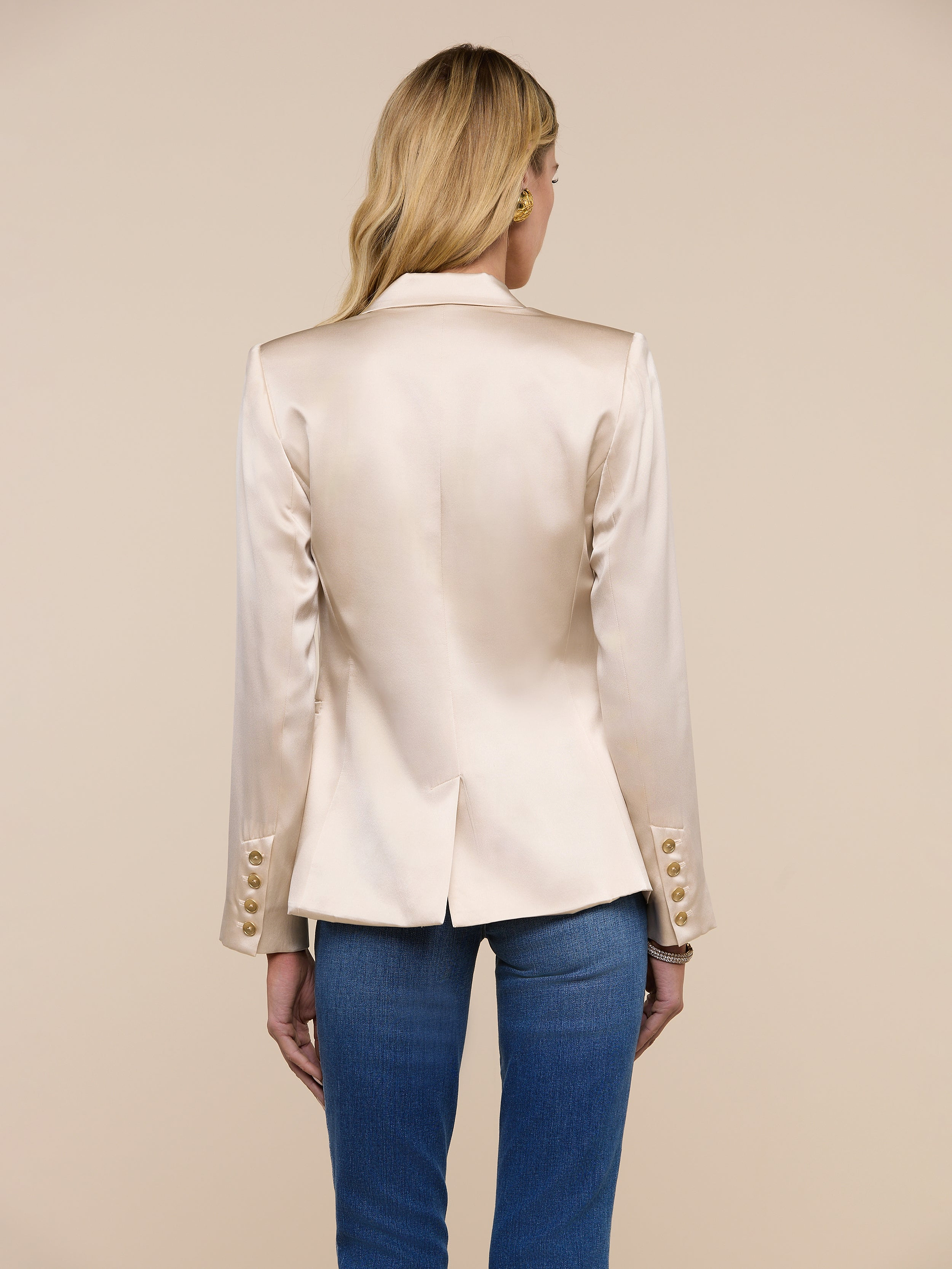 A person with long blonde hair, wearing the Colin Silk Blazer in champagne—featuring buttoned cuffs—paired with blue jeans, stands with their back to the camera against a plain light background.