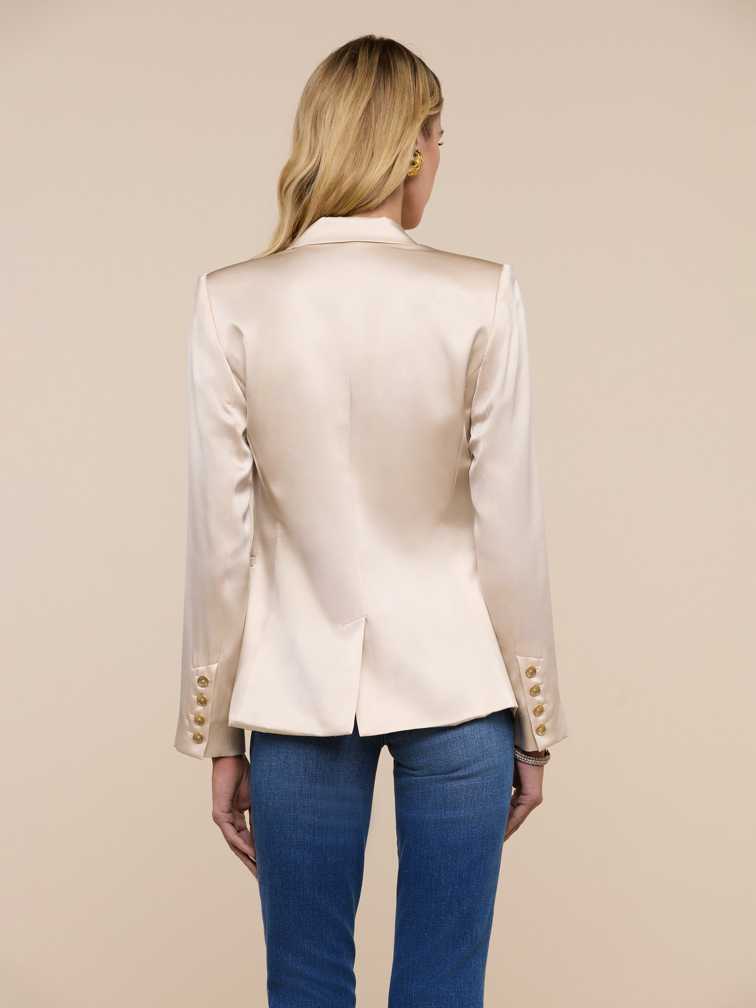 A person with long blonde hair, wearing the Colin Silk Blazer in champagne—featuring buttoned cuffs—paired with blue jeans, stands with their back to the camera against a plain light background.