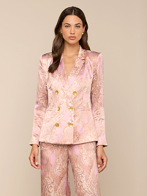 A woman with long brown hair and minimal makeup stands against a beige background wearing the Colin Silk Blazer in shiny pink and gold paisley print, featuring gold buttons.