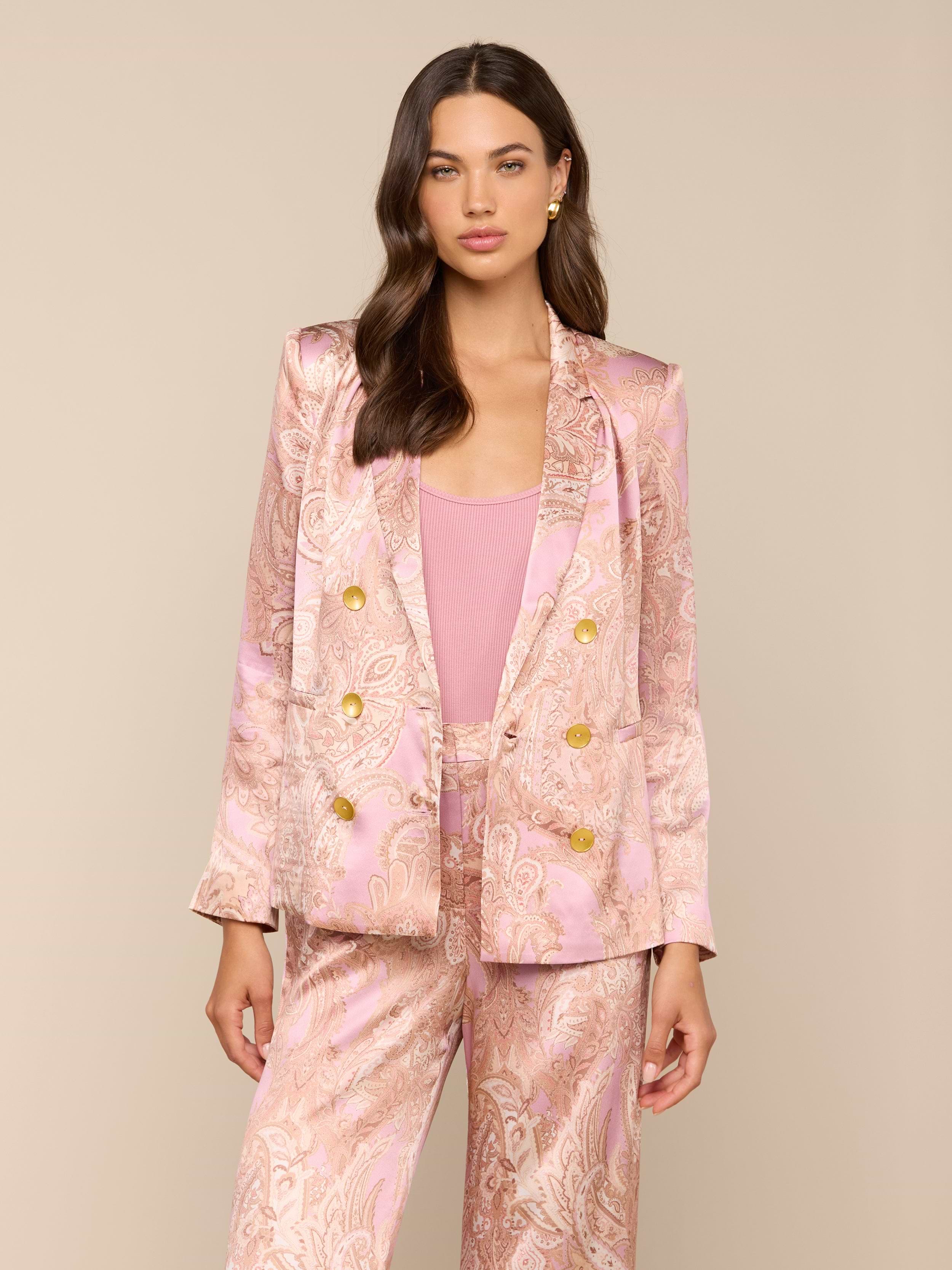 A woman with long brown hair and a neutral expression stands against a plain light background, wearing the Colin Silk Blazer in a pink paisley print with gold buttons.