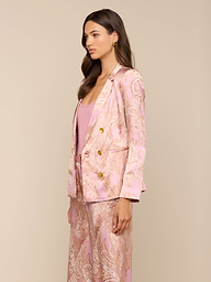 A woman stands in profile against a plain background, wearing the Colin Silk Blazer in a light pink paisley print with a matching top. She has long, wavy brown hair and a neutral facial expression.