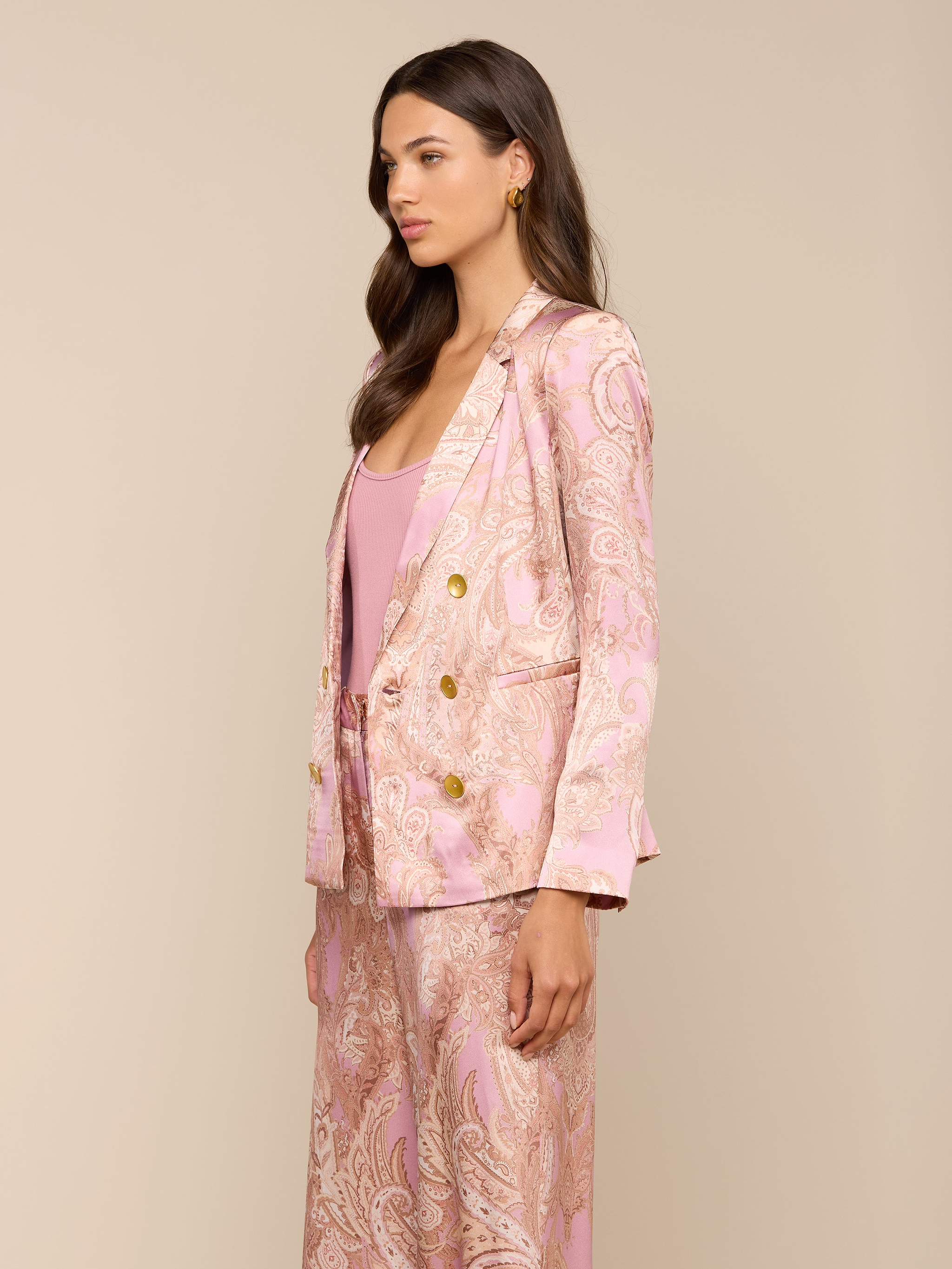 A woman stands in profile against a plain background, wearing the Colin Silk Blazer in a light pink paisley print with a matching top. She has long, wavy brown hair and a neutral facial expression.