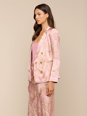 A woman stands in profile wearing the Colin Silk Blazer in pink and gold paisley, paired with matching pants and a light pink silk top. Her long brown hair is styled in loose waves against a beige background.