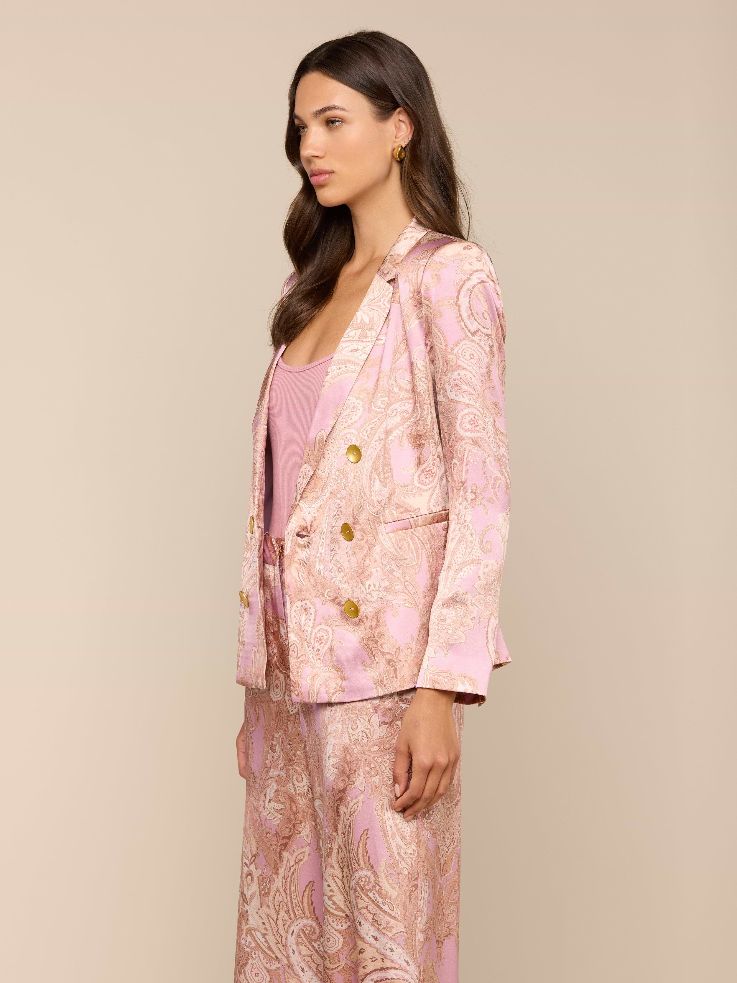 A woman stands in profile against a plain background, wearing the Colin Silk Blazer in a light pink paisley print with a matching top. She has long, wavy brown hair and a neutral facial expression.