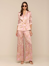 A woman wearing sunglasses and a pink paisley print Colin Silk Blazer with gold buttons and wide-leg pants stands against a plain white background.
