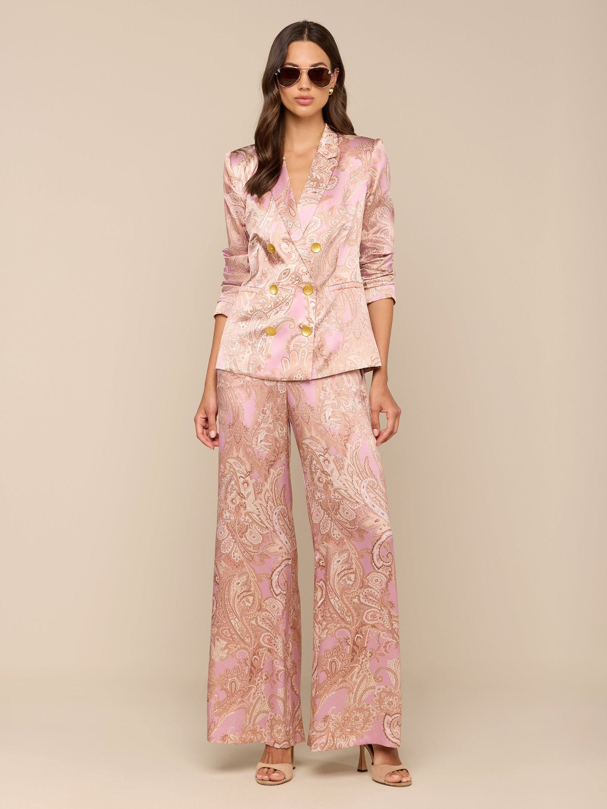 A woman wearing sunglasses and a pink paisley print Colin Silk Blazer with gold buttons and wide-leg pants stands against a plain white background.