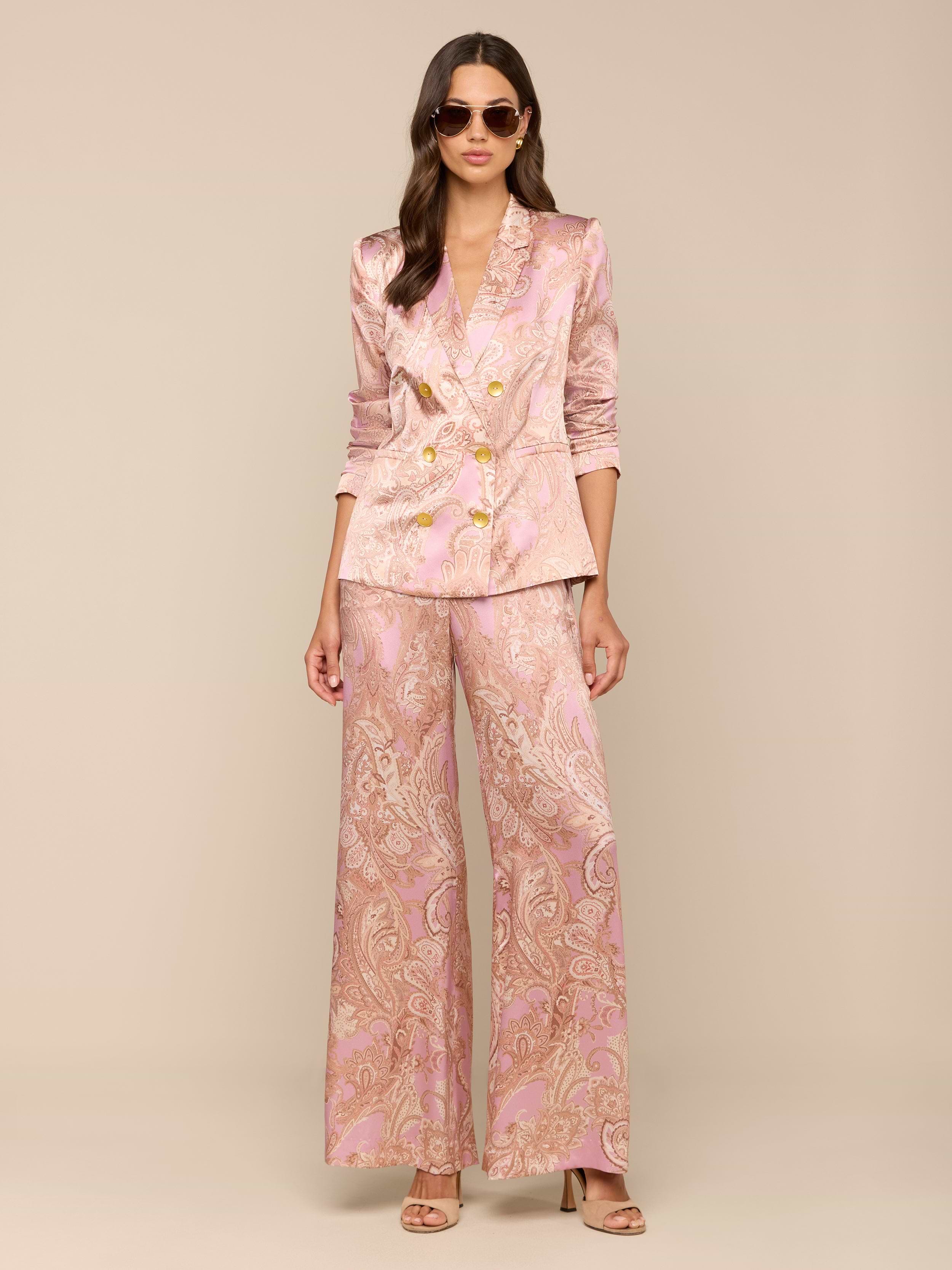 A woman wearing sunglasses and a pink paisley print Colin Silk Blazer with gold buttons and wide-leg pants stands against a plain white background.