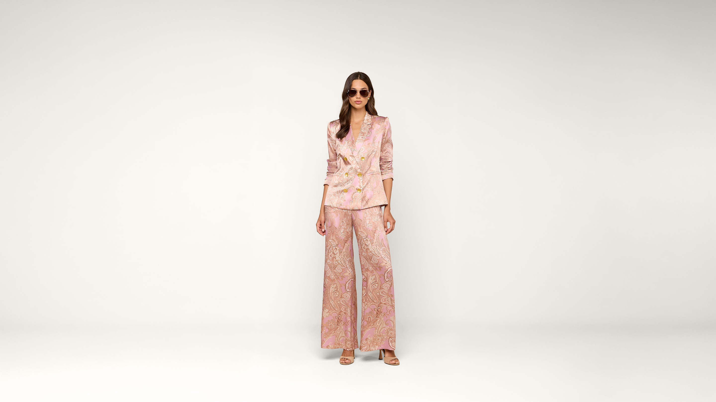 Woman stands against a plain white background wearing sunglasses and a light pink paisley pantsuit with wide-leg pants and a matching blazer. She has long hair and is wearing heels.