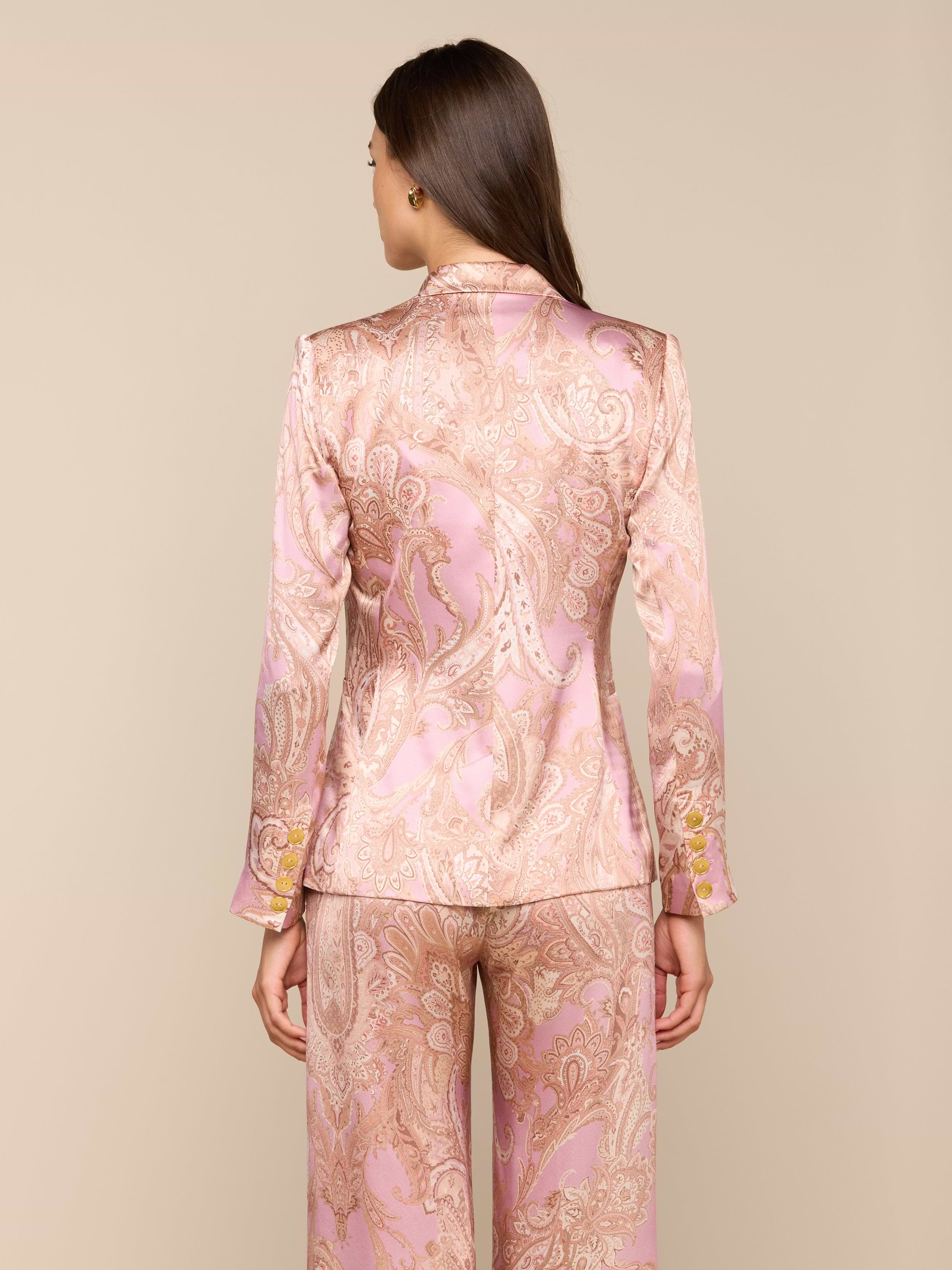 A woman stands facing away, wearing the Colin Silk Blazer and wide-leg pants in a pink and gold paisley print silk charmeuse with long sleeves, set against a plain light background.