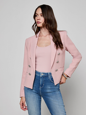 A woman with long brown hair models the Brooke Open-Front Blazer in pale pink over a matching top and high-waisted blue jeans, standing against a plain light gray background with one hand on her hip.