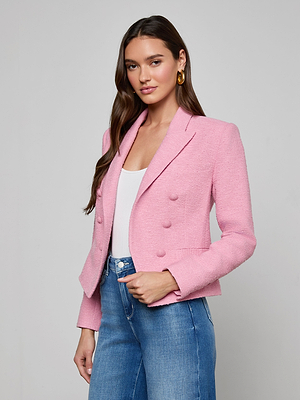 A woman with long brown hair wears the Brooke Tweed Blazer—a pink cotton-blend, double-breasted jacket—over a white top and blue jeans, standing against a plain light gray background.