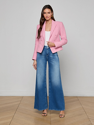 A woman with long brown hair poses indoors against a light wall, wearing the Brooke Tweed Blazer in pink over a white top, paired with wide-leg blue jeans and open-toe heels on a wooden floor.