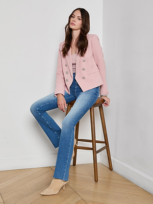 A woman with long brown hair sits on a stool against a white wall, wearing the Brooke Open-Front Blazer in pale pink over a light pink ribbed top, blue jeans, and beige pointed-toe heels, radiating relaxed confidence.
