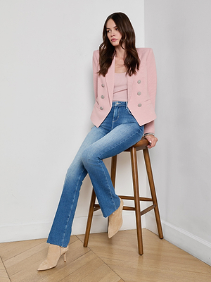 A woman in a pale pink Brooke Open-Front Blazer, light pink ribbed top, blue high-waisted jeans, and beige heeled boots sits on a wooden stool against a white wall and wooden floor.