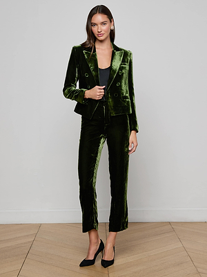 A woman with long brown hair and a neutral expression wears the L'AGENCE Brooke Velvet Blazer in dark green, styled with matching pants, a black top, and black heels, standing against a plain background with one hand by her side.