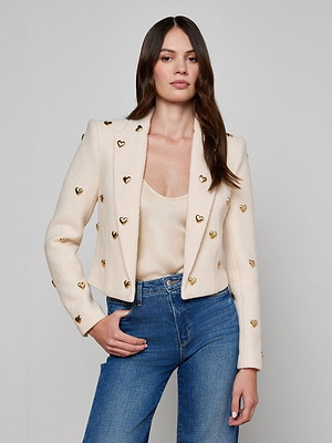A woman with long brown hair wears the Jen Embellished Blazer—cream-colored and adorned with gold heart accents—over a matching top and blue jeans, standing against a plain light gray background.