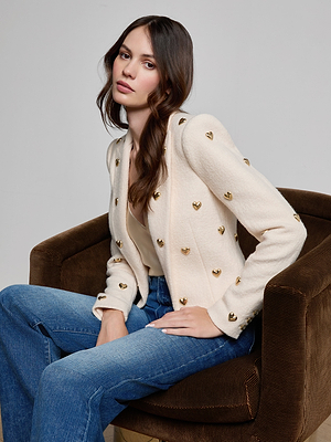 A woman with long brown hair sits on a brown chair, wearing the Jen Embellished Blazer—a cream cropped blazer with gold heart accents—paired with a light top and blue jeans, looking at the camera with a neutral expression.