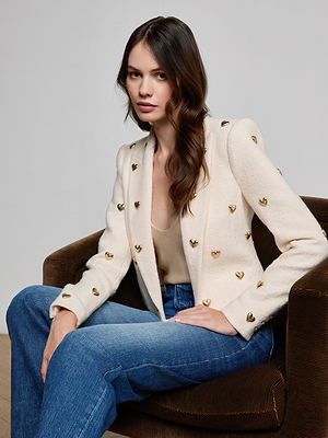 A woman with long brown hair sits on a brown chair, wearing the Jen Embellished Blazer over a beige top and blue jeans. She looks toward the camera with a neutral expression.