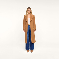 A woman stands against a plain background, facing forward with her arms at her sides, wearing the Kolina Long Coat in Italian wool cognac brown over a light top, paired with blue wide-leg jeans and brown shoes.