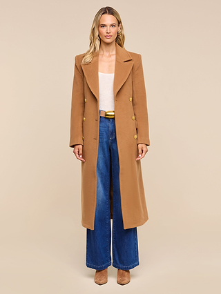 A woman stands facing forward, wearing the Kolina Long Coat—a long Italian wool coat in a camel hue and true power coat—paired with wide-leg blue jeans, a white top, and brown shoes, against a plain beige background.