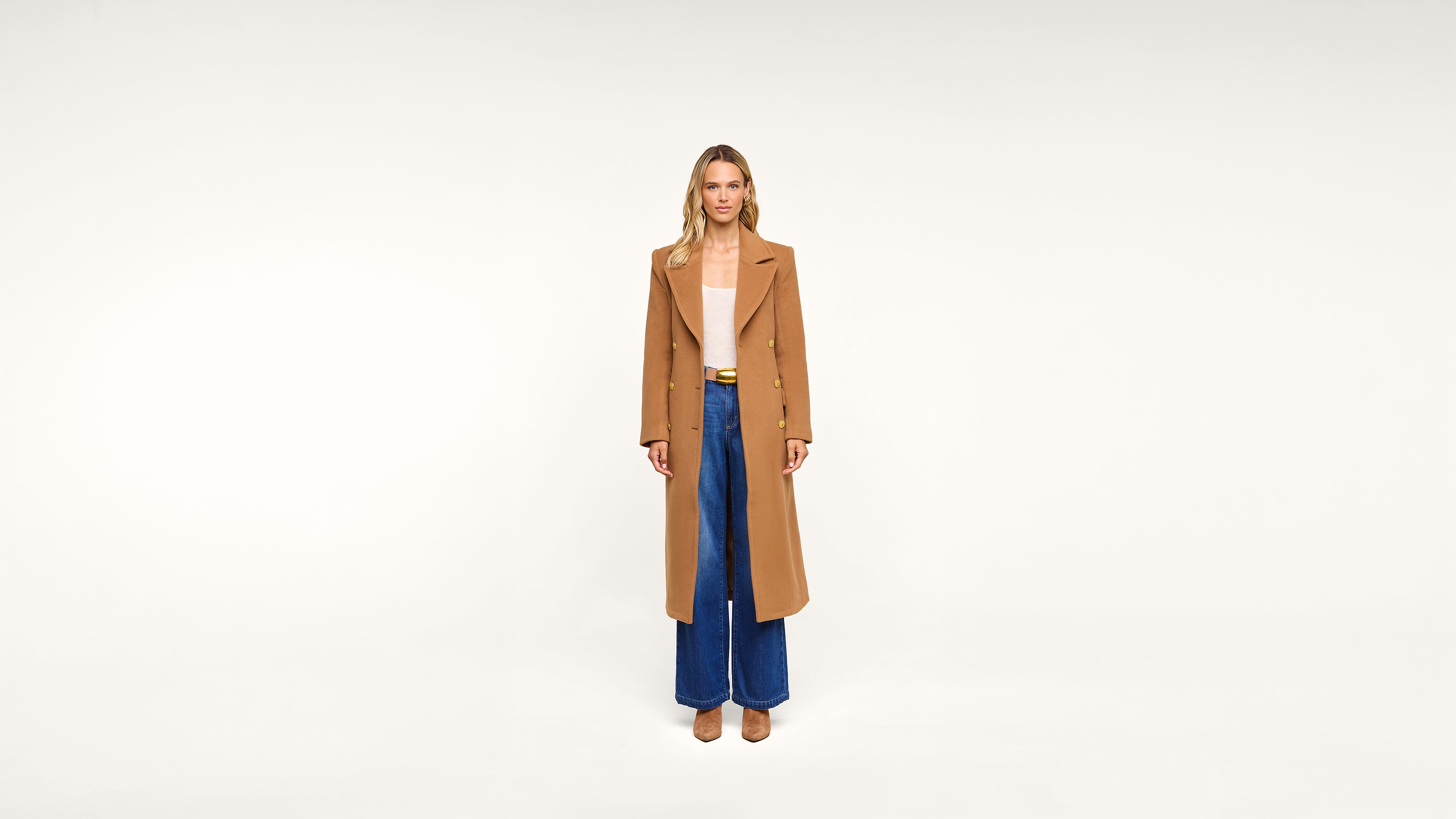 A woman stands facing forward, wearing a tan long coat, light top, blue wide-leg jeans, and tan shoes, against a plain white background.