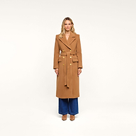A woman stands facing forward wearing the Kolina Long Coat, a long Italian wool coat in camel featuring gold buttons, wide lapels, and a belt. She pairs it with wide-leg blue jeans and tan shoes, set against a plain white background.