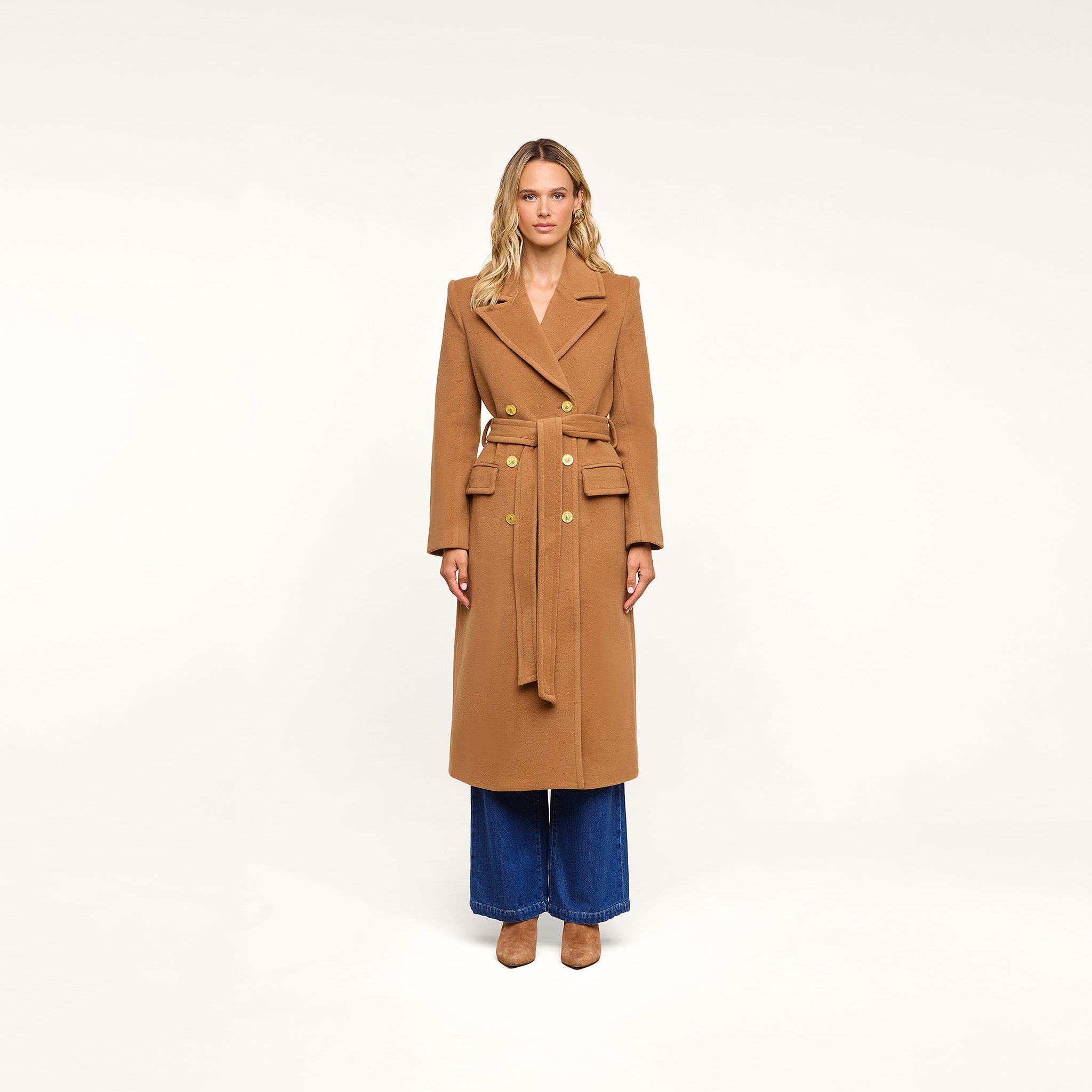 A woman stands facing forward wearing the Kolina Long Coat, a long Italian wool coat in camel featuring gold buttons, wide lapels, and a belt. She pairs it with wide-leg blue jeans and tan shoes, set against a plain white background.