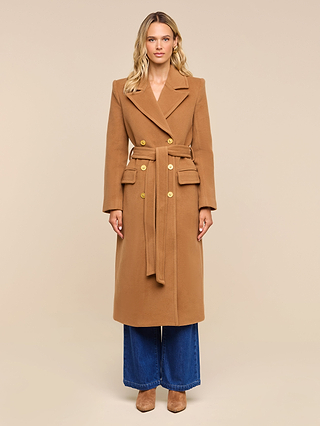 A woman stands facing forward, wearing the Kolina Long Coat—a long camel-colored Italian wool power coat with a belt—paired with wide-leg blue jeans and tan shoes, against a plain beige background.
