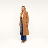 A woman stands against a plain background, wearing the Kolina Long Coat in camel over a light top, paired with blue wide-leg jeans and brown shoes.