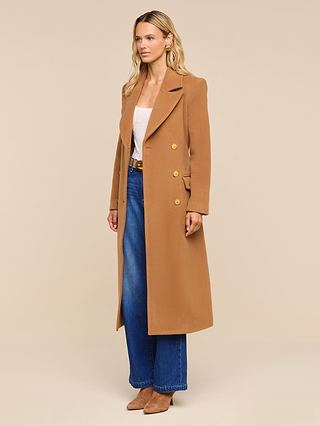 Wearing the Kolina Long Coat in camel over a light top, blue wide-leg jeans, and brown heeled boots, a woman stands against a plain beige background.