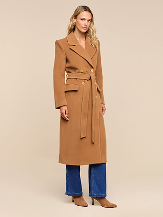 A woman wearing the Kolina Long Coat in Italian wool camel, paired with blue jeans and cognac brown heeled boots, stands against a beige background.