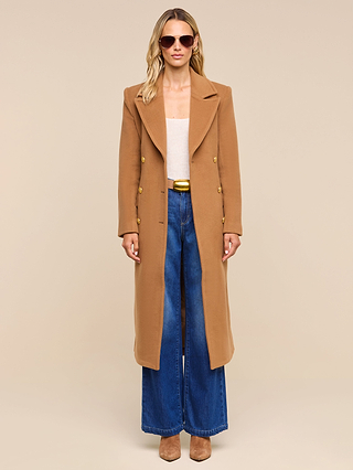 A person wearing sunglasses, the Kolina Long Coat in camel, a light top, wide-leg blue jeans, and cognac brown shoes stands against a beige background.