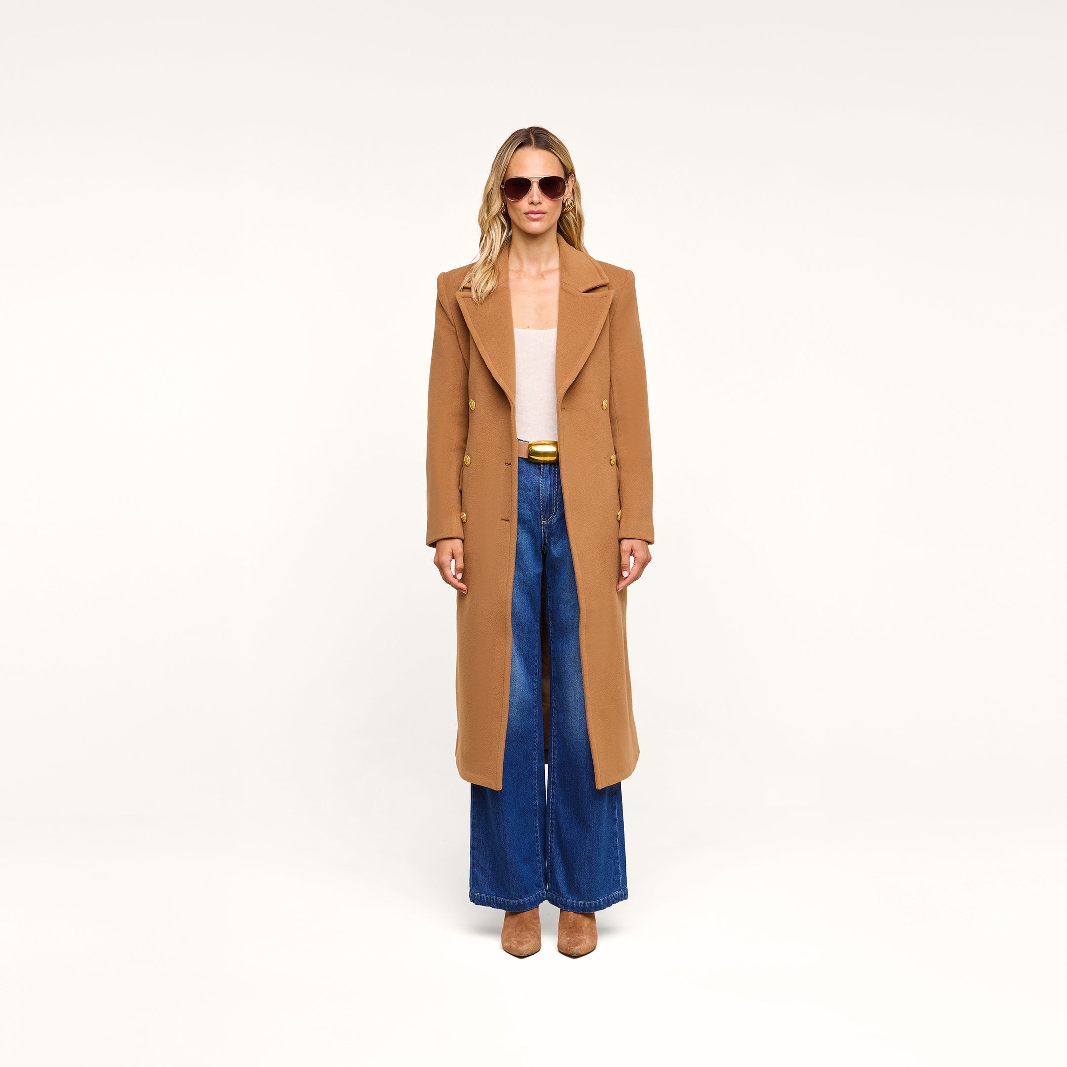 A woman wearing sunglasses, the Kolina Long Coat in camel, a beige top, wide-leg blue jeans, and tan shoes stands against a plain white background.