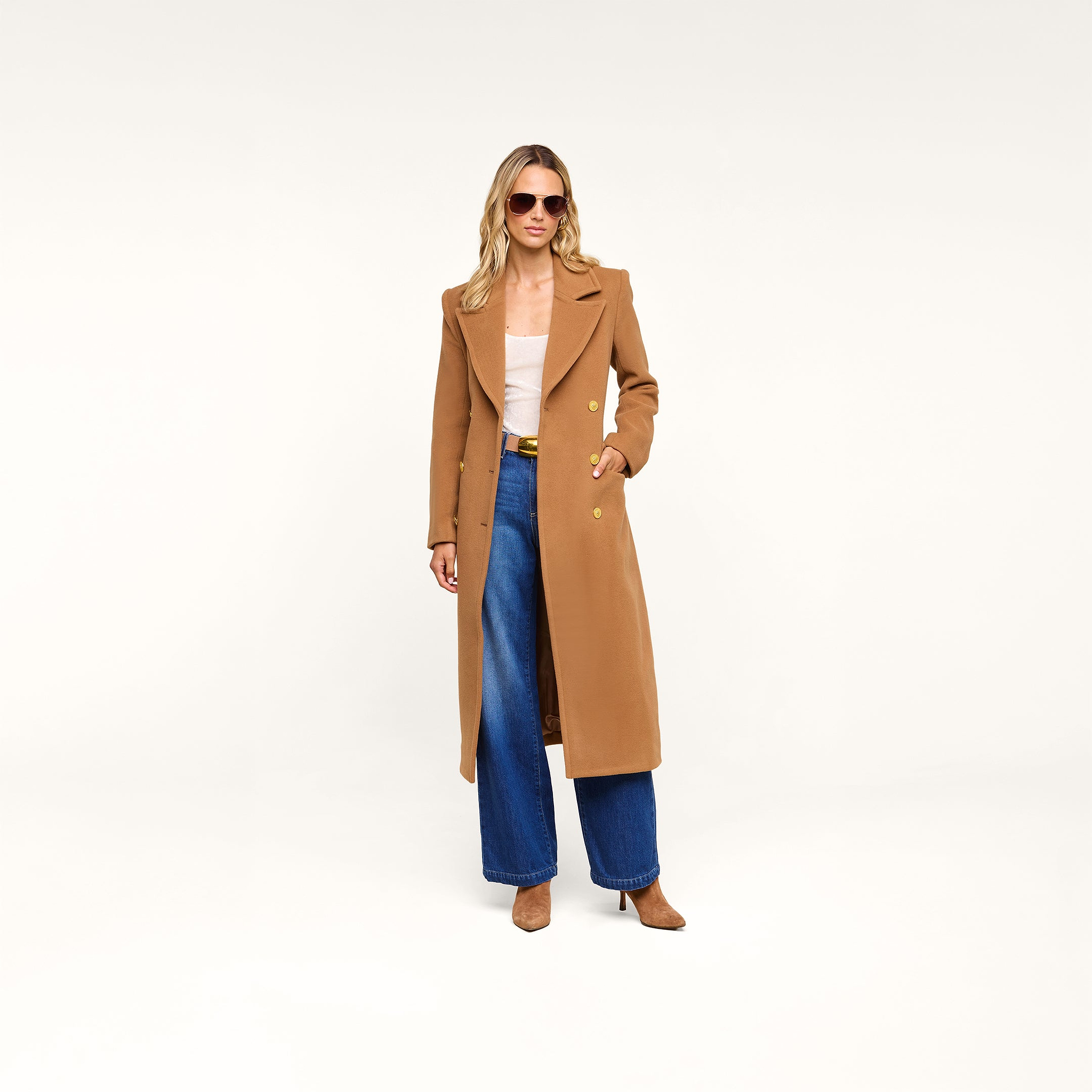 Wearing the Kolina Long Coat in cognac brown, a woman stands confidently against a plain white background. She completes her look with sunglasses, wide-leg blue jeans, a beige top, and brown heels.