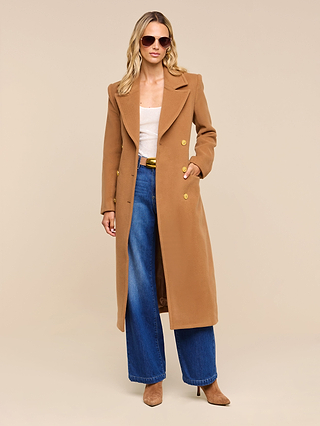 A woman wearing sunglasses, the Kolina Long Coat in cognac brown, a white top, blue wide-leg jeans, and tan heeled boots stands against a beige background.