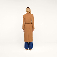 A person with long blonde hair stands facing away, wearing the Kolina Long Coat in camel-colored Italian wool with a belt, paired with wide-legged blue jeans, against a plain white background.