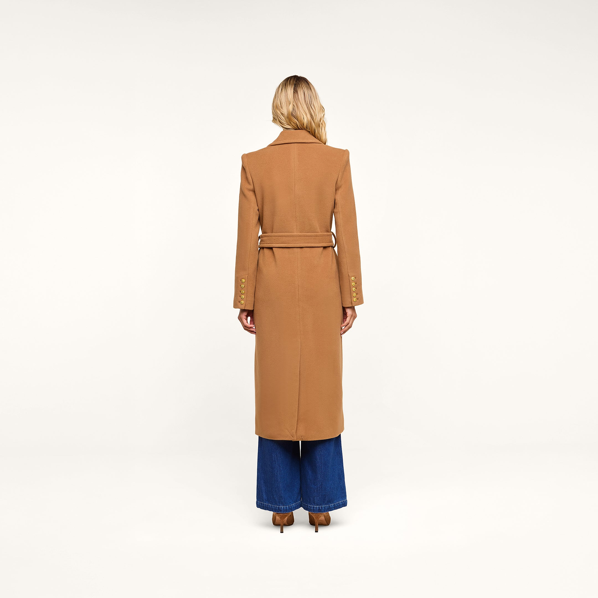 A person with long blonde hair stands facing away, wearing the Kolina Long Coat in camel-colored Italian wool with a belt, paired with wide-legged blue jeans, against a plain white background.