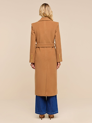 Wearing the Kolina Long Coat—a long, camel-colored Italian wool coat with a belt and gold buttons—a person with long blonde hair, seen from behind, pairs it with blue wide-leg jeans and tan heels, standing against a plain beige background.