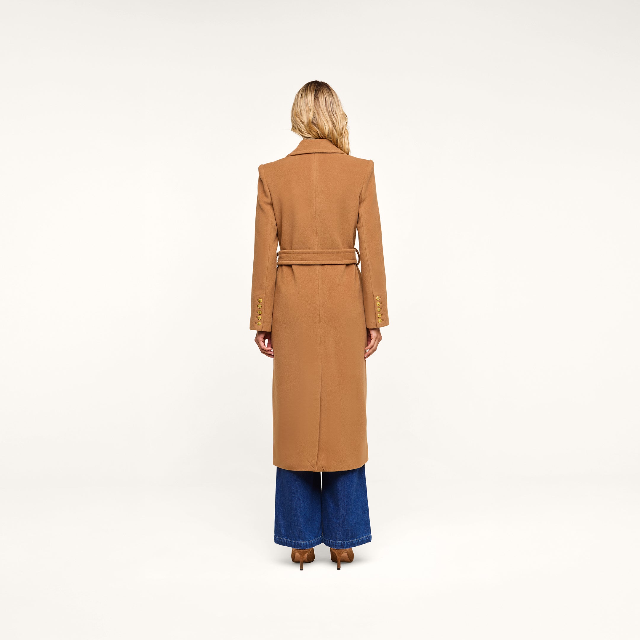 A person with long blonde hair stands facing away, wearing the Kolina Long Coat in camel-colored Italian wool with a belt, paired with wide-legged blue jeans, against a plain white background.