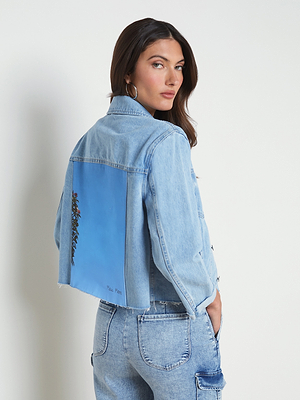 A woman with long brown hair wears a light blue cropped denim jacket featuring a painted blue panel with floral detail on the back, paired with high-waisted blue jeans. She stands sideways, looking over her shoulder.