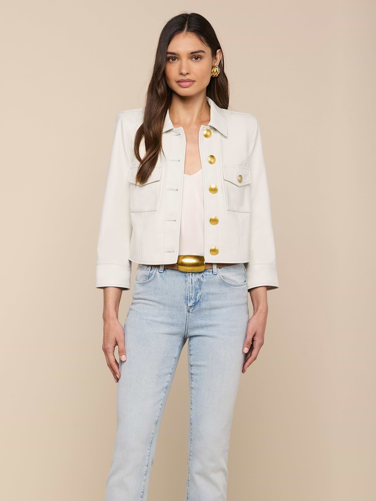 Donna con lunghi capelli castani che indossa la giacca di pelle Kumi Cropped Leather Jacket bianca con bottoni dorati, un top bianco, jeans blu chiaro e orecchini dorati, in piedi su uno sfondo beige.