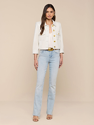 A woman stands against a plain white background, wearing the Kumi Cropped Leather Jacket with gold buttons, a white top, light blue jeans, and beige heeled sandals. She has long brown hair and a neutral expression.