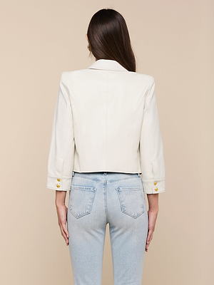A woman with long brown hair is seen from the back, wearing the Kumi Cropped Leather Jacket in white with gold sleeve buttons and light blue high-waisted jeans, set against a beige background.