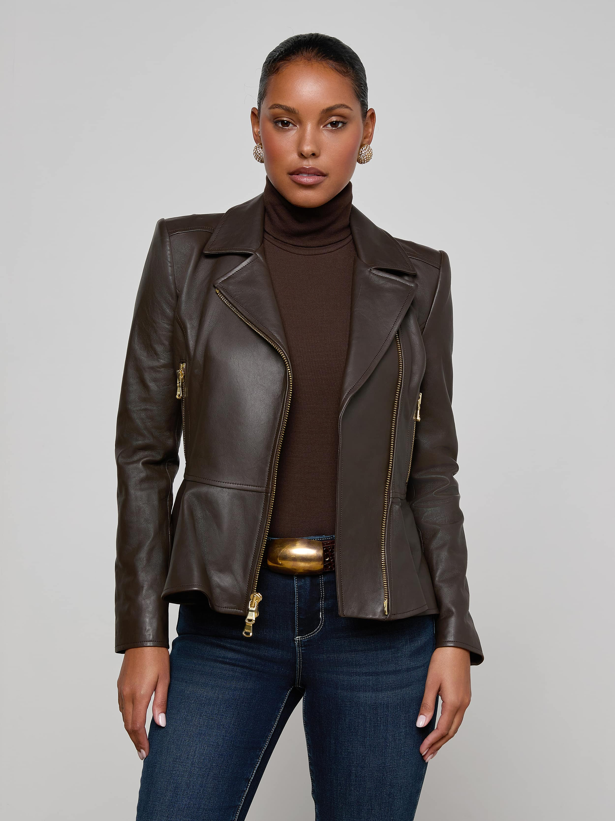A woman wearing the Lyric Leather Peplum Jacket in dark brown over a brown turtleneck and blue jeans, standing against a plain light background.